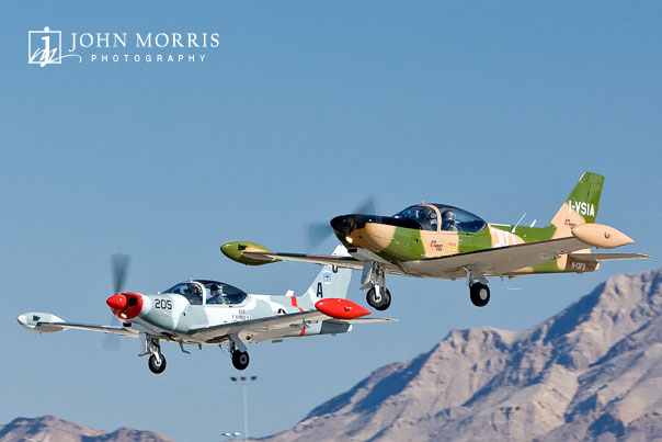 Two fighter training aircraft fly low and in formation over a desert air strip.