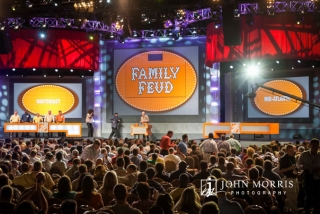 A capacity crowd of attendees pack a convention hall to watch and participate in a mock Family Feud entertainment segment of a corporate event.