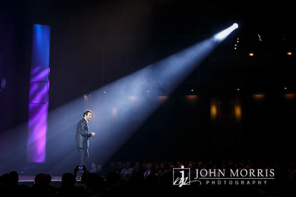 An executive delivers a keynote speech to his audience and is dramatically spotlighted, on stage, from a single light source