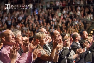 A capacity crowd of attendees, dress in business attire, giving a standing ovation for a speaker during a corporate event.