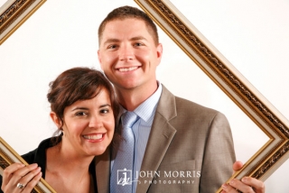 A happy couple poses in front of a white background and behind an empty frame in a photo booth portrait during a corporate event.