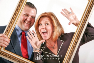 A happy couple poses in front of a white background and behind an empty frame in a photo booth portrait during a corporate event.