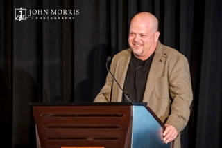 Pawn star, Rick Harrison, smiles from behind a podium while giving a presentation during a corporate event.