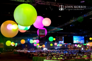 Giant, colorful globes, suspended from the cieling of a massive arena provide a fun and elegant atmosphere above a thousand exquisitely decorated tables in anticipation of a gala corporate event.