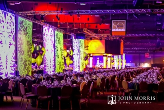 A massive, elaborated decorated arena is filled with thousands of exquisitely decorate tables in anticipation of a gala event honoring attendees during a corporate event.
