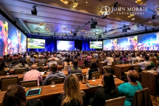 A crowded conference room, full of attendees, surrounded in a dazzling display of big screens, listen intently to a presentation during a corporate event.