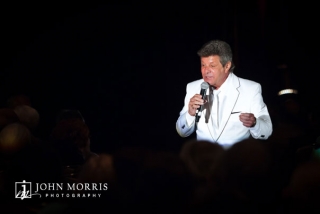 Frankie Avalon is isolated on stage in dramatic fashion from a single spotlight as he performs for an audience during a convention.