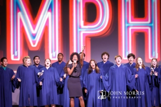 A choir, on stage, joyously belts out background vocals as an energetic singer raises her hand towards a crowd of attendees during a convention
