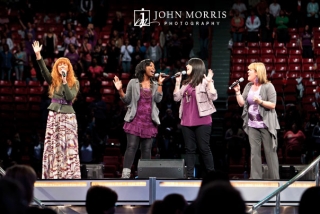 Four women, members of a vocal group, passionately perform a song on stage for large audience during a conference.