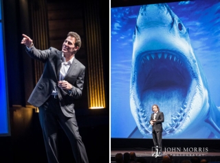 A giant image of a great white shark, projected on a massive screen behind a speaker, creates a comical but strong impression on the audience during a speech at a conference.