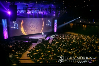 The spotlight shines on a CEO as he delivers his keynote speech to a crowded arena as seen from the upper levels.