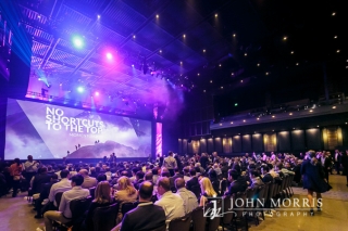 Wide angle view of a brilliantly lit stage and venue filled to capacity with attendees during a keynote speech.