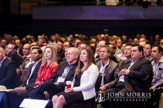 Audience of attendees in professional business attire listening inventively to a keynote speaker during a conference.