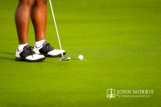 A closeup of a golfers shoes, and his putter head as he strikes a golf ball on an immaculate putting surface during a corporate outing.