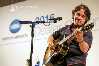 John Oates, on stage, playing guitar for an intimate crowd of attendees during a corporate event.