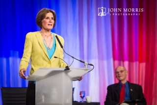 Mary Matalin, on stage addressing a Corporate Event with husband James Carville looking on