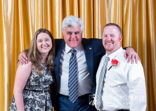 Comedian Jay Leno hugging a couple during a meet & greet in front of a gold backdrop.