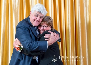 Comedian Jay Leno hugging a fan during a meet & greet in front of a gold backdrop.